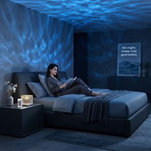 A woman relaxing in a modern dark bedroom illuminated by calming ocean wave light projections from a bedside lamp.