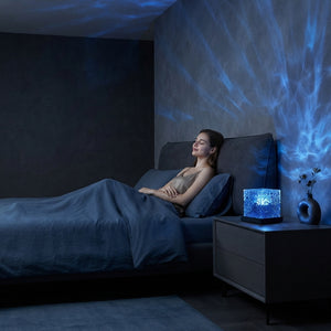 A woman relaxes in a dark, modern bedroom filled with calming blue ocean wave light projections from a bedside lamp.