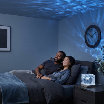 A couple relaxing in a modern, dim bedroom illuminated by blue ocean wave patterns projected from a lamp on a bedside table.